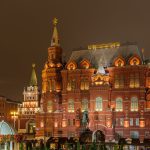 Красное здание Исторического музея с подсвеченными башнями вечером, перед ним памятник маршалу Жукову и осенние клумбы