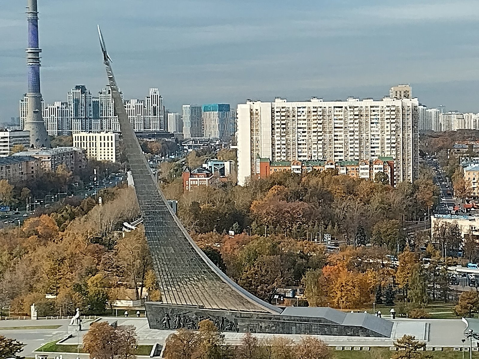 Титановый монумент «Покорителям космоса» с ракетой на вершине среди осенних деревьев, вдали Останкинская башня