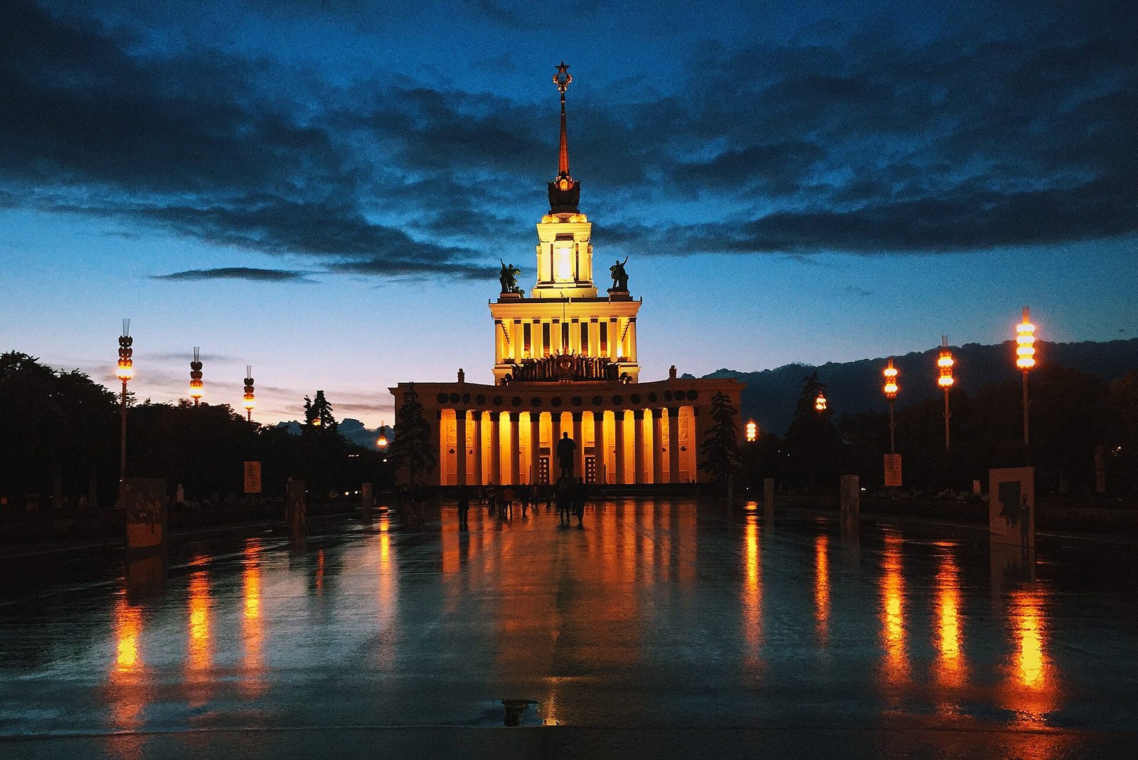 Подсвеченный Центральный павильон ВДНХ со шпилем и фонари вдоль главной аллеи вечером