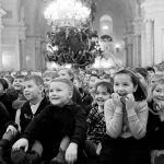 Улыбающиеся дети в зале Большого Кремлёвского дворца смотрят новогоднее представление, за ними нарядная ёлка под люстрой