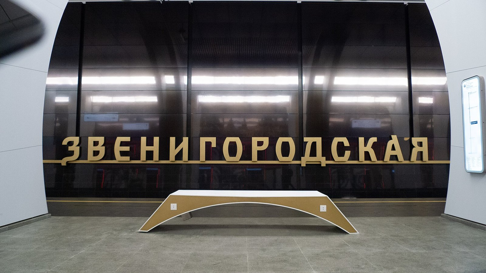 Золотистые буквы названия «Звенигородская» на тёмной путевой стене и светлая изогнутая скамья на платформе