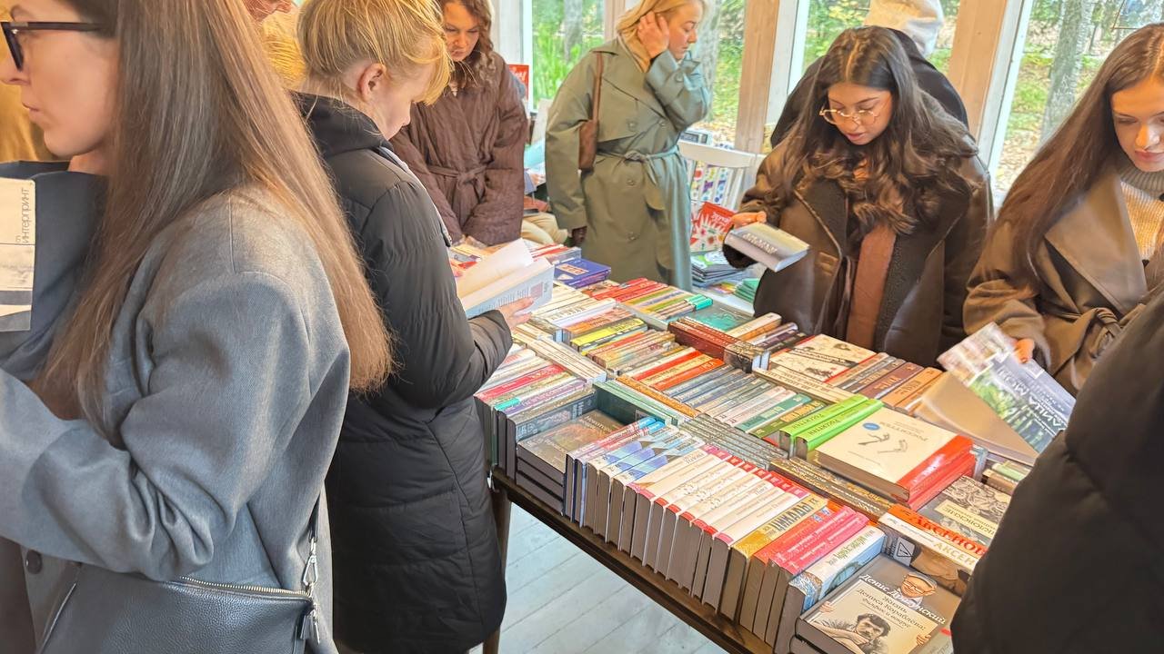 Длинный стол с рядами книг в светлом зале с большими окнами, посетители листают книги